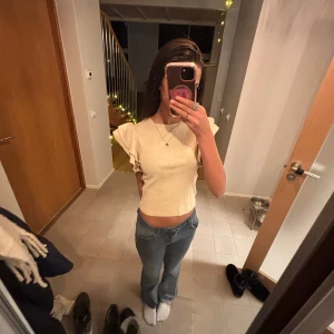 Beige ribbad topp med volangärmar från  - Säljer en snygg beige ribbad topp med volangdetaljer på ärmarna. Toppen är croppad och har en tight passform, perfekt för en trendig look. 