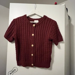 Vinröd kofta från H&M - Säljer en vinröd kofta från H&M med korta ärmar och snygga knappar framtill. Koftan är ribbad och har en mysig känsla, perfekt för höst och vinter. Passar bra till både jeans och kjol.