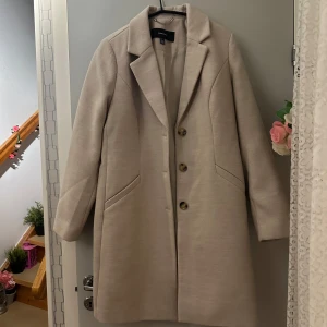 Beige kappa från Vero Moda - Elegant beige kappa från Vero Moda med klassisk design och knappar framtill. Perfekt för en stilren look under kyligare dagar. Kappan har långa ärmar och två fickor för extra funktionalitet.