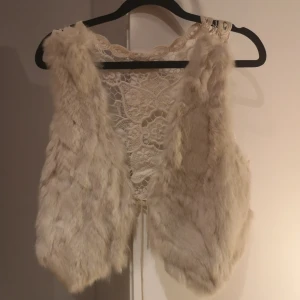 Vit pälsväst med spetsdetaljer från Culture - Säljer en unik vit pälsväst från Culture med vackra spetsdetaljer på ryggen. Västen har en bohemisk känsla och är perfekt för att ge din outfit en extra touch. Den är fodrad med ett mönstrat tyg och har en knytning framtill. Passar perfekt för höst och vår.