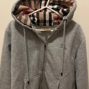 Grå zip hoodie från Burberry - Säljer en stilren grå hoodie från Burberry med dragkedja och klassiskt rutigt mönster i huvan. Tillverkad i mjuk bomull för extra komfort. Perfekt för en avslappnad och trendig look.