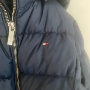 Blå dunjacka från Tommy Hilfiger - Säljer en snygg blå dunjacka från Tommy Hilfiger med dragkedja och huva. Jackan har det klassiska Hilfiger-emblemet på bröstet och är perfekt för kyliga dagar. Den är både stilren och funktionell.