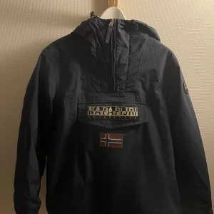 Mörkblå jacka från Napapijri - Säljer en mörkblå jacka från Napapijri med huva och dragkedja. Jackan har en stor ficka framtill med Napapijri-logga och en norsk flagga. Insidan är fodrad med grått tyg för extra värme. Hör gärna av dig vid minsta lilla fundering 💯