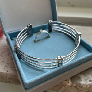 Silverfärgat armband och ring - Elegant set med ett silverfärgat armband och matchande ring. Armbandet har en stilren design med flera parallella band och små dekorativa detaljer. Perfekt för att ge en sofistikerad touch till din outfit.