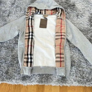 Grå Burberry hoddie  - Säljer en stilren grå huvtröja från Burberry med klassiskt rutigt mönster på insidan av huvan. Tröjan har dragkedja framtill och ribbade muddar vid ärmslut och nederkant.  Hör av er vid frågor