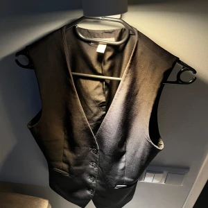 Svart polyester väst från H&M - Snygg svart väst från H&M med knappar framtill. Perfekt för en stilren look. Tillverkad i polyester med en liten del elastan för extra komfort och elegans. Perfekt julklapp 