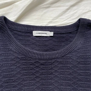  J.Lindeberg knitted sweater - Snygg mörkblå stickad tröja från J.Lindeberg med ett diskret mönster. Perfekt för kyligare dagar med sin klassiska design och bekväma passform. Tröjan har långa ärmar och ribbade kanter. Sitter bra på mig som bär M-L