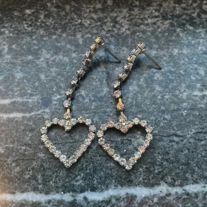 Glittriga hjärtformade örhängen - Snygga örhängen med hjärtformad design och glittriga stenar. Perfekta för att ge en extra touch av glamour till din outfit. De har en elegant och modern stil som passar till många tillfällen.