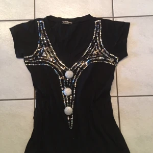 Svart topp med paljetter och knappar - Snygg svart topp med glittrande paljetter och stora dekorativa knappar framtill. Toppen har en v-ringad design och korta ärmar, perfekt för en festlig kväll.