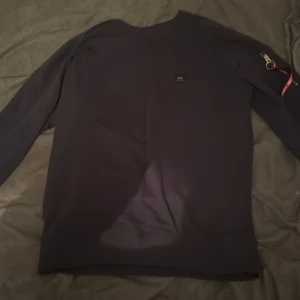 Svart tröja med dragkedja och tag från Alpha Industries - Säljer en svart långärmad tröja med en cool 'Remove Before Flight'-tag. Tröjan har en dragkedja på ärmen och ribbade muddar vid ärmslut och nederkant. Perfekt för en avslappnad stil.