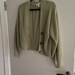 Olivgrön stickad kofta från H&M - Säljer en olivgrön stickad kofta från H&M med stora knappar framtill. Koftan har en avslappnad passform och långa ärmar, perfekt för kyliga dagar. Den är mjuk och bekväm att bära.