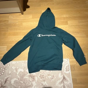 Grön hoodie från Champion - Säljer en grön hoodie från Champion med stor logga på ryggen. Den har en klassisk känguruficka och en bekväm huva. Perfekt för en avslappnad stil.