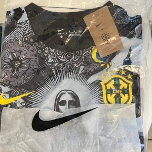 Custom Brazil fotbollströja från Nike - Snygg fotbollströja från Nike med ett unikt mönster i svart och grått. Tröjan har korta ärmar och pryds av Brasiliens emblem samt en stor grafisk design av Kristusstatyn. Perfekt för fotbollsfans som vill sticka ut.