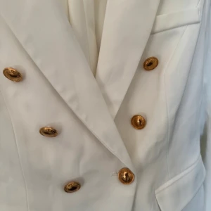 Vit kavaj med guldknappar - Elegant vit kavaj med dubbelknäppning och stiliga guldknappar. Perfekt för en sofistikerad look. Kavajen har en klassisk passform med fickor framtill.