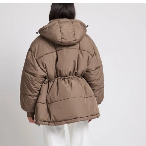 Brun pufferjacka med huva - Säljer en brun pufferjacka med huva och dragkedja. Jackan har en oversized passform och långa ärmar, perfekt för kyliga dagar. Den har även knappar och elastisk midja för extra komfort.