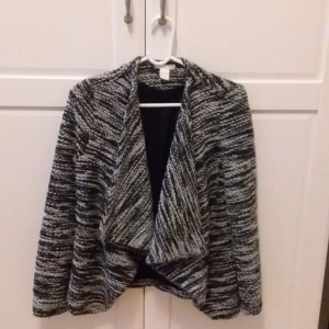 Svartvit stickad jacka - Säljer en mysig svartvit stickad jacka med ett melerat mönster. Jackan har en öppen design utan knappar och långa ärmar. Perfekt för kyliga dagar.