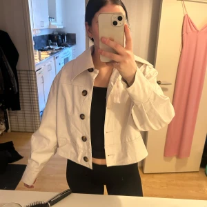 Vit jacka från Gina Tricot - Säljer en stilren vit skinn jacka från Gina Tricot i storlek XS. Jackan har stora knappar och en croppad passform! Använd ett par gånger!