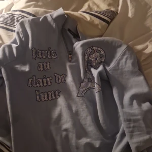 Ljusblå tröja med text och motiv - Säljer en ljusblå tröja med texten 'Paris au clair de lune' och ett motiv av Eiffeltornet i glittriga detaljer. Tröjan har långa ärmar och en bekväm passform. Perfekt för en avslappnad stil med en touch av Paris.