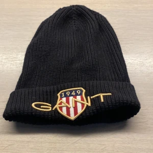 Svart ribbad mössa från GANT - Snygg svart ribbad mössa från GANT med broderad logga och emblem framtill. Perfekt för kyliga dagar och en stilren look.