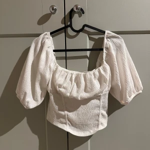 Vit blus/topp från Gina Tricot - Säljer en söt vit blus från Gina Tricot med puffiga ärmar och en fyrkantig urringning. Tyget har en lätt textur som ger en fin detalj till toppen. Bra pris!