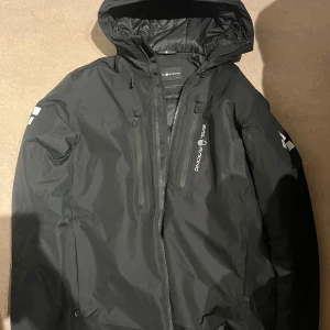 Svart jacka från Sail Racing - Säljer en svart jacka från Sail Racing med huva och dragkedja. Jackan är vattentät, vindtät och andas bra. Den har flera praktiska fickor med dragkedjor och är perfekt för utomhusaktiviteter.