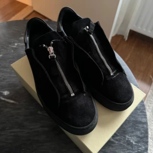 Axel Arigato Svarta  med dragkedja - Snygga svarta sneakers med dragkedja framtill. Skorna har en stilren design med en rund tå och är tillverkade i mocka. Perfekta för en modern och avslappnad stil.