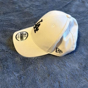 Beige keps från LA NEW ERA - Snygg beige  keps från LA NEW ERA med justerbar rem baktill. Den har en svart broderad logotyp på framsidan och är perfekt för en avslappnad stil.
