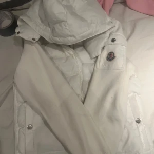 Vit pufferjacka från Moncler - Säljer en snygg vit pufferjacka från Moncler med avtagbar huva och dragkedja. Jackan har långa ärmar och tryckknappar på fickorna. Perfekt för kyliga dagar!