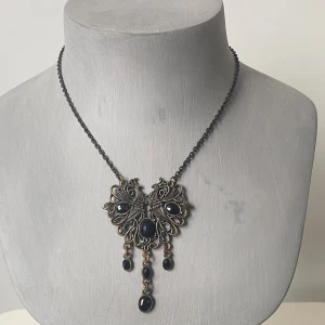 Vintageinspirerat halsband med svarta stenar - Säljer ett supercoolt vintageinspirerat halsband med en unik design. Halsbandet har en mörk kedja och ett stort hänge  med svarta stenar. Detaljerna är riktigt snygga och ger en elegant touch. Perfekt för att piffa upp en outfit 