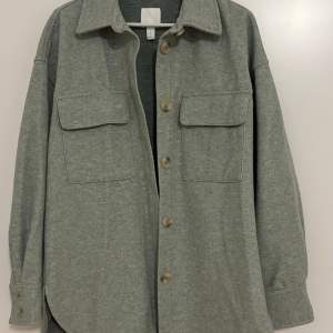 Säljer en stilren grå overshirt med knappar framtill. Den har två stora bröstfickor och långa ärmar. Perfekt för lager-på-lager under hösten eller våren. Skön och enkel att matcha med andra plagg.