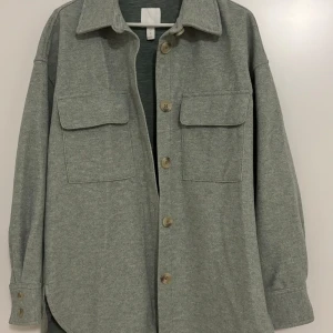 Grå overshirt med knappar - Säljer en stilren grå overshirt med knappar framtill. Den har två stora bröstfickor och långa ärmar. Perfekt för lager-på-lager under hösten eller våren. Skön och enkel att matcha med andra plagg.