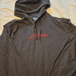 Svart hoodie från Hollister - Säljer en svart hoodie från Hollister med röd broderad logga på bröstet. Den är i storlek XS och har en skön passform med långa ärmar och en praktisk ficka framtill. Perfekt för höst och vinter när du vill vara både bekväm och stilren. 🖤