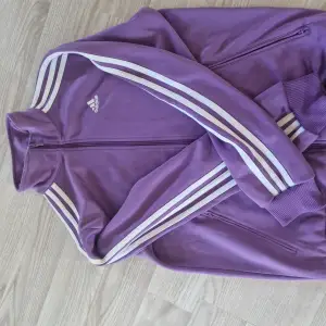 Säljer en snygg lila träningsjacka från Adidas. Den har de klassiska tre vita ränderna längs ärmarna och en dragkedja framtill. Perfekt för träning eller som en sportig vardagslook. Jackan är i bra skick och redo för nya äventyr!