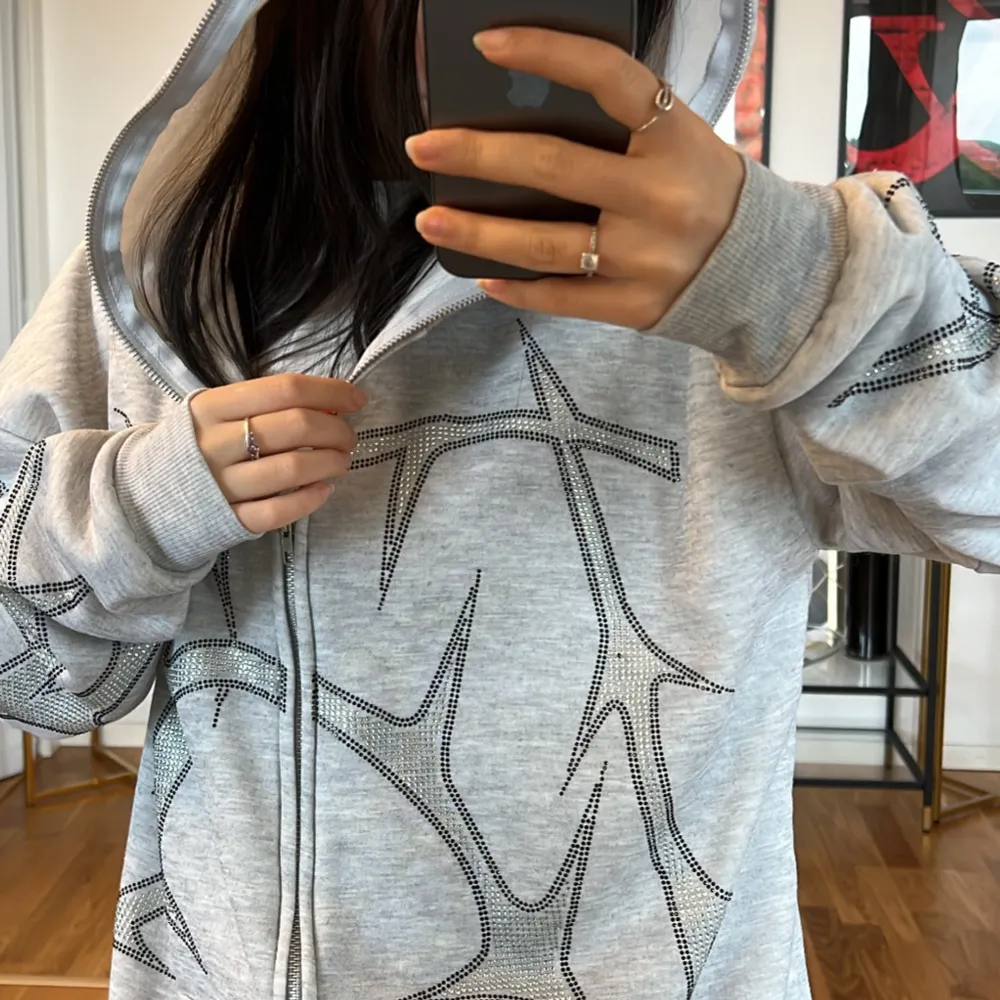 Säljer en supermysig grå hoodie med ett coolt svart mönster. Den har en dragkedja framtill och en stor huva som är perfekt för kyliga dagar. Tröjan är oversized och gjord i mjukt material, vilket gör den perfekt för både vardag och chill. Den har långa ärmar och ribbade muddar vid ärmslut och nederkant. Perfekt för höst och vinter!. Tröjor & Koftor.