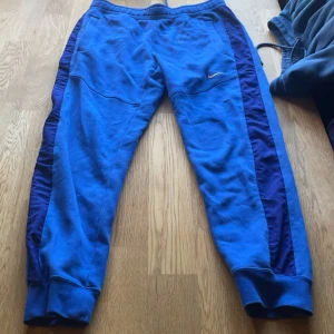 Blå mjukisbyxor från Nike - Säljer ett par blå mjukisbyxor från Nike. De har en avslappnad passform med resår i midjan och muddar vid bensluten. Byxorna har en snygg kontrast med mörkare blå ränder längs sidorna och en liten Nike-logga på framsidan. Perfekta för chill eller träning!