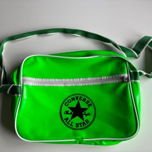Grön axelväska från Converse - Säljer en grön axelväska från Converse All Star. Väskan har en stor svart logga på framsidan och en praktisk dragkedja. Den är perfekt för vardagsbruk och har en justerbar axelrem. Väskan är i syntetmaterial och har vita detaljer