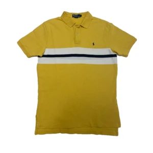 Polo Ralph Lauren piké gul - Säljer en snygg gul pikétröja från Ralph Lauren. Den har korta ärmar och en klassisk krage med knappar. Tröjan har en vit och mörkblå rand över bröstet och det ikoniska broderade logotypen på framsidan. Perfekt för en avslappnad men stilren look!