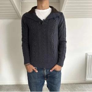 Mörkblå stickad tröja med knappar - Säljer en snygg mörkblå stickad tröja med kabelstickat mönster. Tröjan har en unik krage med knappar och bruna armbågslappar som ger en cool touch. Perfekt för höst och vinter, och den är supermysig att ha på sig. Passar perfekt till jeans för en avslappnad look.
