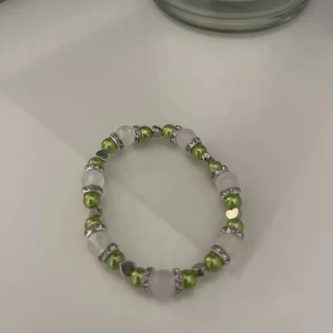 Grönt och vitt pärlarmband - Snyggt armband med gröna och vita pärlor, perfekt för att piffa upp vilken outfit som helst. Armbandet har också små silverdetaljer som ger det en extra touch av elegans. Det är elastiskt och passar de flesta handleder.
