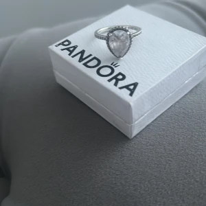 Silverring från Pandora med stor sten - Elegant silverring från Pandora med en stor, droppformad klar sten i centrum. Ringen har en glittrig design med små stenar längs bandet. Perfekt för att ge en lyxig touch till din stil.