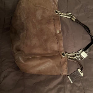 Brun skinnväska från Jimmy Choo - Snygg brun skinnväska från Jimmy Choo med robusta metallspännen och detaljer. Perfekt för att bära dina dagliga nödvändigheter med stil. Väskan har en rymlig design och en elegant look.