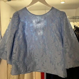 Blå spetsblus med rosett - Säljer en elegant blå spetsblus med vackra rosettdetaljer på baksidan. Blusen har trekvartsärmar och ett blommigt mönster som ger en romantisk känsla. Från neo noir storlek 40