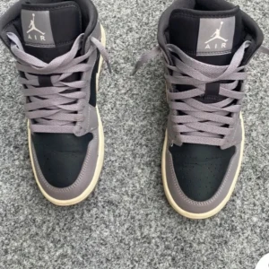 Nike Air Jordans 1 - Snygga Nike Air Jordans i en stilren kombination av grått och svart. Skorna har en klassisk design med snörning och en vit sula som ger en fräsch kontrast. Perfekta för dig som vill ha både stil och komfort.
