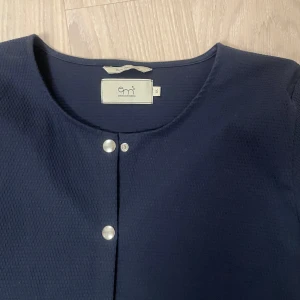 Mörkblå kofta från Emma och Malena  - Säljer en stilren mörkblå kofta från Emma och Malena med rund halsringning och knappar framtill. Koftan har långa ärmar och en enkel design som passar perfekt till olika tillfällen. Perfekt för att hålla sig varm och snygg!