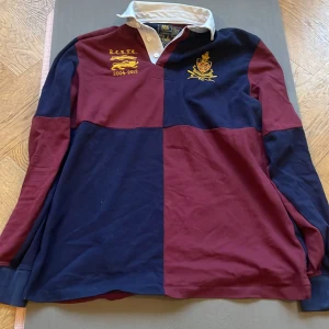 Randig pikétröja från Polo Ralph Lauren - Säljer en snygg randig pikétröja från Polo Ralph Lauren i marinblått och vinrött. Tröjan har långa ärmar och en klassisk krage med knappar. Broderade detaljer i gult på bröstet ger en exklusiv touch. Perfekt för en stilren look!