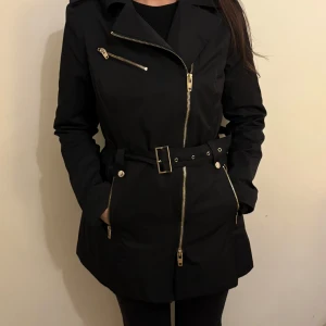 Svart trenchcoat med bälte - Snygg svart trenchcoat från Rock and blue med dragkedja och bälte i midjan. Jackan har långa ärmar och flera fickor med dragkedjor. Perfekt för en stilren look. Har liten skada som enkelt går att sy igen.