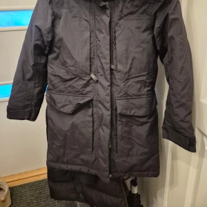 Svart parkajacka från Didriksons - Säljer en svart parkajacka från Didriksons med fuskpäls på huvan och dragkedja framtill. Jackan har långa ärmar och stora fickor, perfekt för kyliga dagar. Insidan är fodrad för extra värme. 