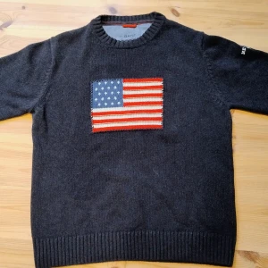 Mörkgrå tröja med amerikansk flagga från Gant - Säljer en snygg mörkgrå tröja från Gant med en broderad amerikansk flagga på framsidan. Tröjan har ribbade muddar och rund halsringning. Perfekt för en casual look!  Tröjan är i storlek: L men är liten i storleken