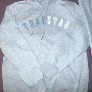 Grå hoodie från Trapstar - Säljer en grå hoodie från Trapstar med text i blått och vitt på bröstet. Den har en klassisk design med dragsko i huvan och ribbade muddar vid ärmslut och nederkant. Perfekt för en avslappnad stil.