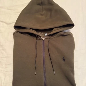 Olivgrön hoodie från Ralph Lauren - Säljer en stilren olivgrön hoodie från Ralph Lauren. Den har en dragkedja framtill och en klassisk broderad logga på bröstet. Lite sliten nere vid dragkedjan, syns knappt och påverkar inte användning. Nypris 2299